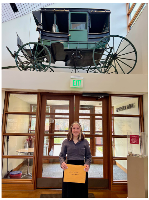 High school student standing in front of a door with an old school vehicle displayed above her